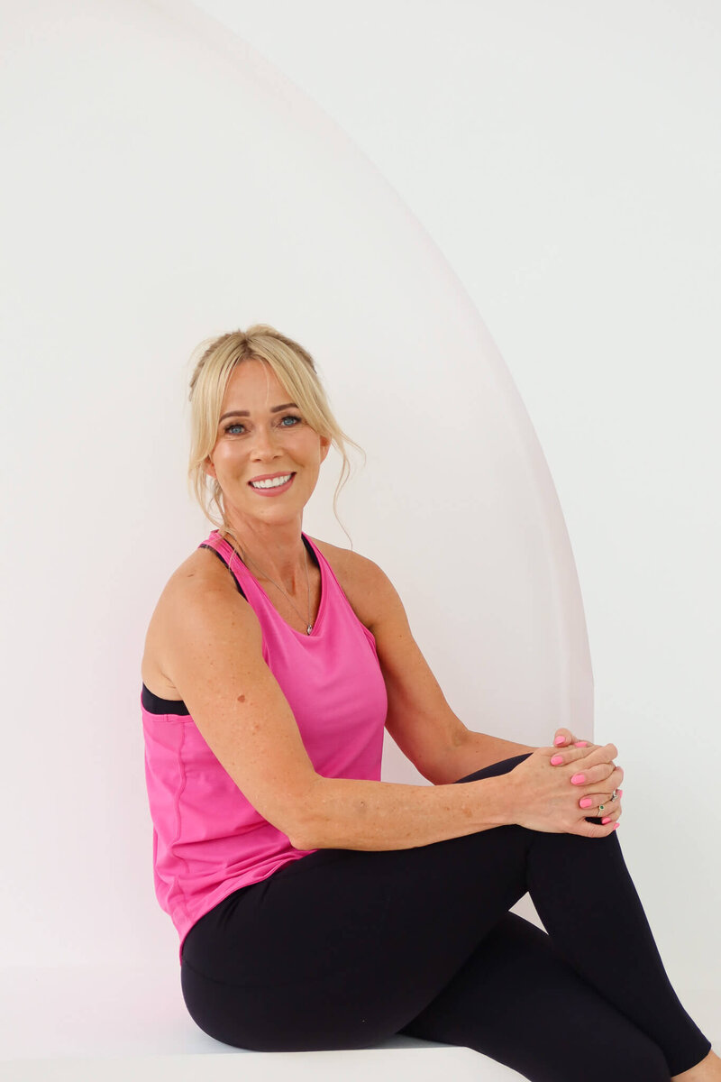 A smiling woman relaxes in a white bathtub, holding a bottle of rosé sparkling wine in one hand and an empty wine glass in the other, with sheer white curtains and a window in the background.