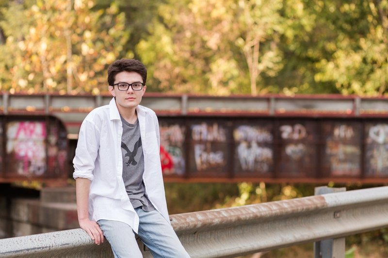 waynedale high school senior sitting  on guard rail with graffetti bridge behind him, serious look on his face, white shirt unbuttoned over a tshirt, photographed by Jamie Lynette Photography Canton Ohio Senior Photographer