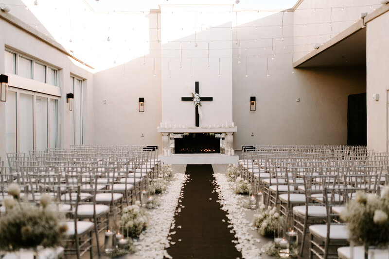 A romantic ceremony aisle at SoHo63, a Chandler wedding and event venue, is lined with white rose petals and floral arrangements leading to a fireplace adorned with a cross. The modern outdoor chapel space is lit with soft string lights, creating a peaceful setting for a meaningful wedding ceremony.
