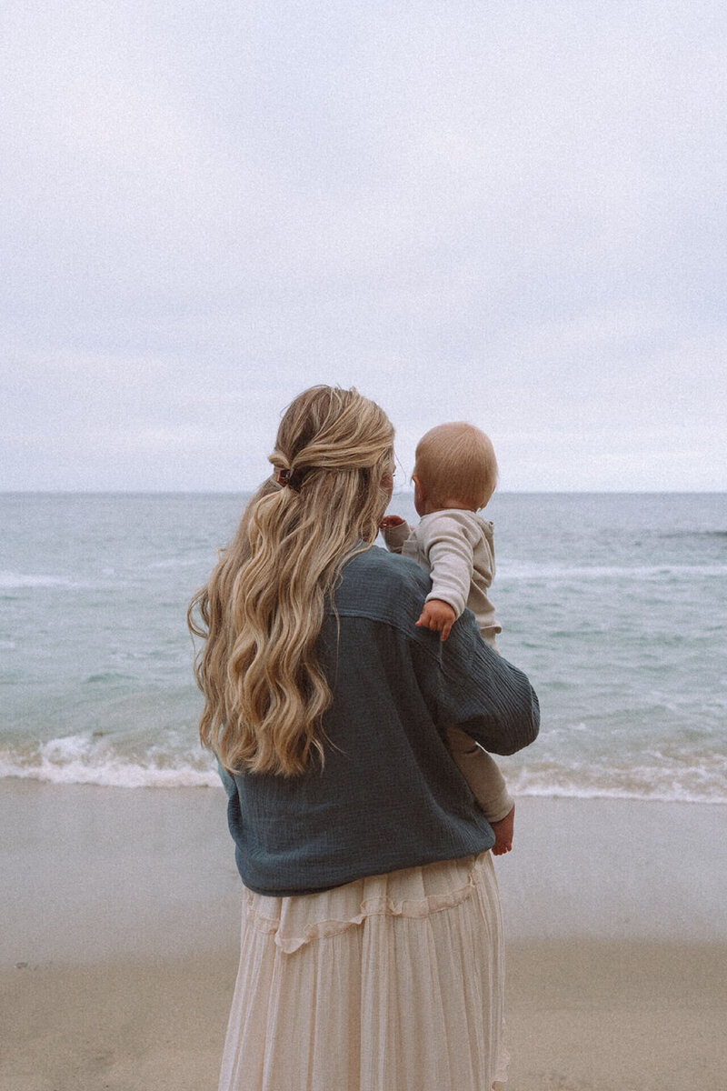 mother watching ocean while holding baby in neutral outfit