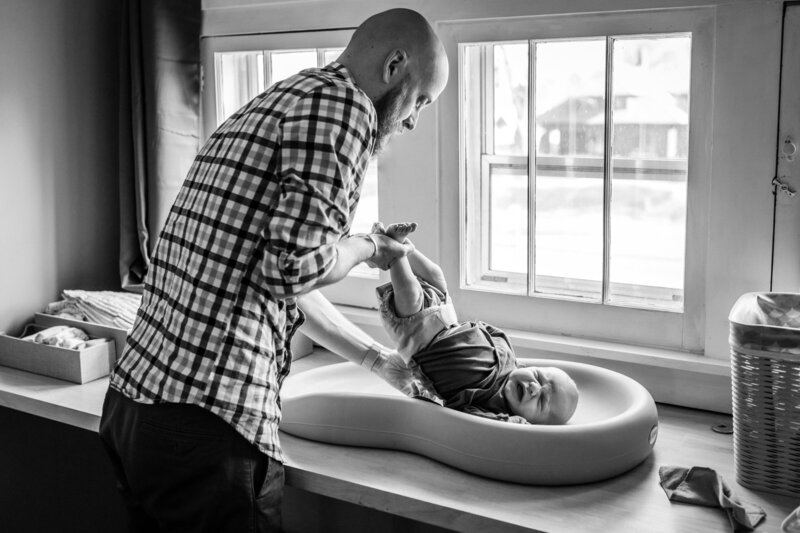 A black and white image of a father changing his baby's diaper on a changing table by a window. The father, dressed in a plaid shirt, is gently holding the baby’s legs, while the baby lays on the changing pad, visibly upset and crying. The room is softly lit by natural light coming through the window.