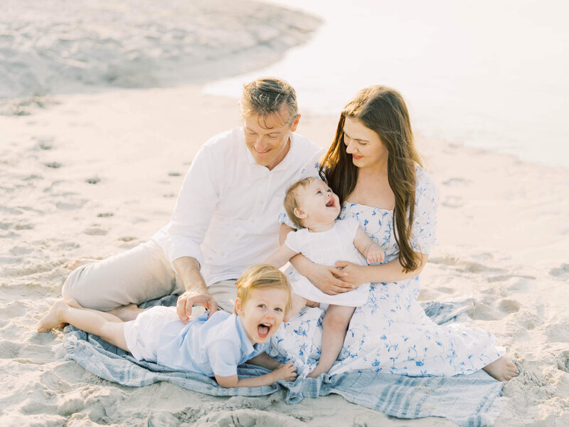 mother and father playing with their two children while sitting on a blanket on the beach dressed in nice clothes.
