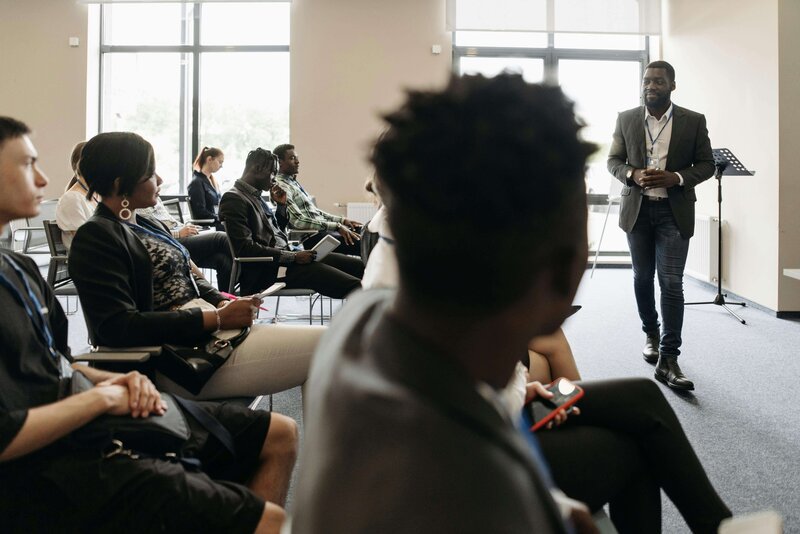 A professional man speaking in front of a group seated in a conference room. The audience is engaged, with some taking notes, while the presenter gestures confidently near a podium.
