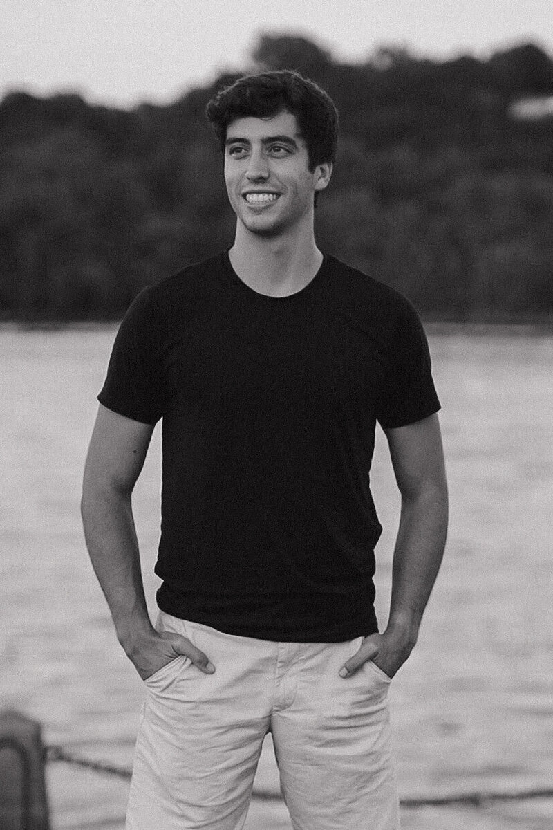 Adam stands with a denim button up shirt and khakis against a white wall. He is holding his videography camera down by his side as he smiles at the camera