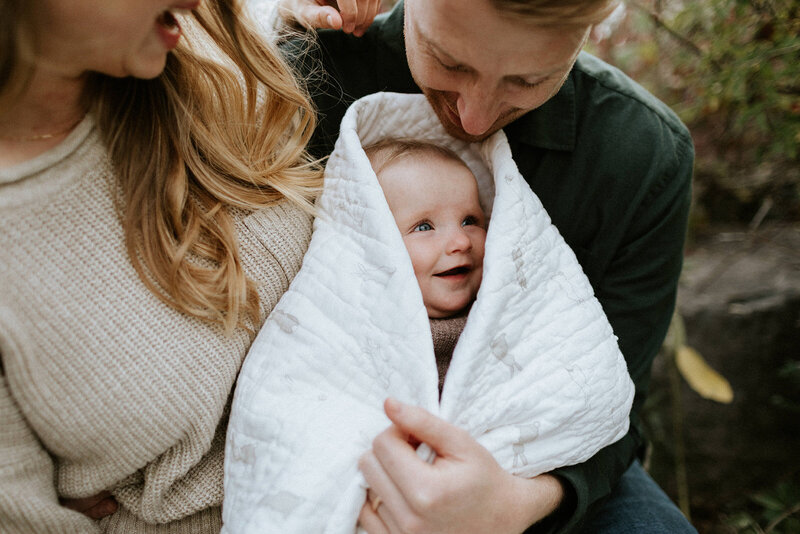 A smiling baby wrapped in a white blanket looks up at an adult holding them, with another person nearby—a perfect moment captured by a family photographer in Toronto.