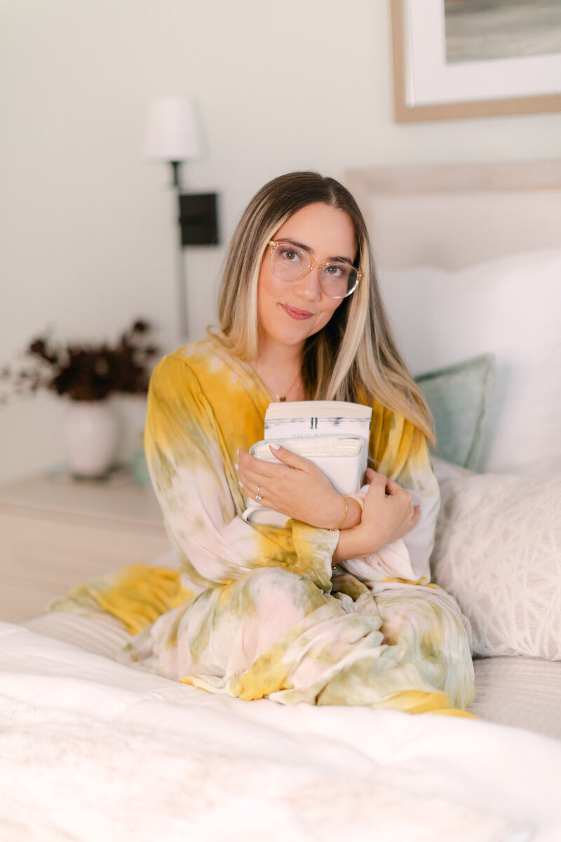 Merlyn Asencio holding a book with a warm smile in bed, embodying a nurturing, calming energy central to her trauma therapy practice in Miami, Florida