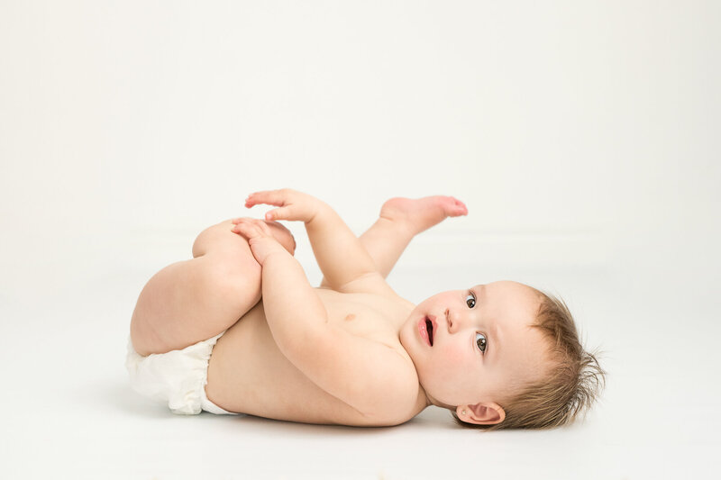 Baby wearing diaper playing with his feet in simple white background