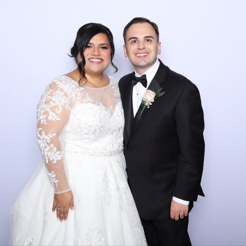 Newlywed couple smiling in front of a light background, bride in a lace wedding gown and groom in a black tuxedo, captured at AR Photobooth.