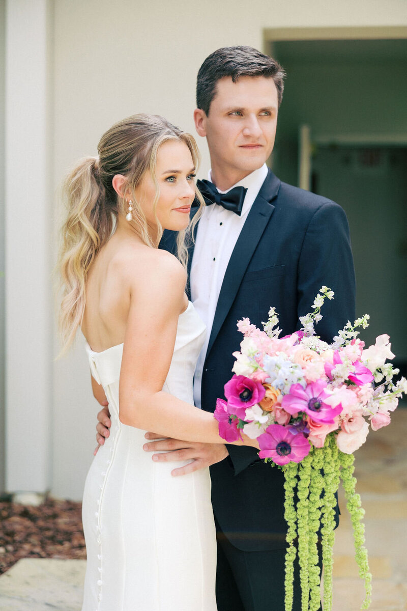 Bride with blonde hair and white dress stands with groom outside the Athens Chapel holding pink and white bouquet