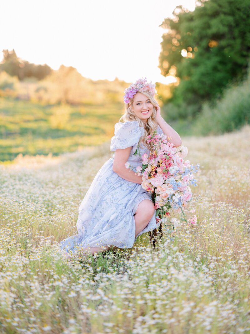 A woman with long soft blonde curls and a floral headpiece wearing a blue floral gown sits in a flower field holding a bouquet of flowers and smiling, Bay area Family photographer Light Livin Photography.
