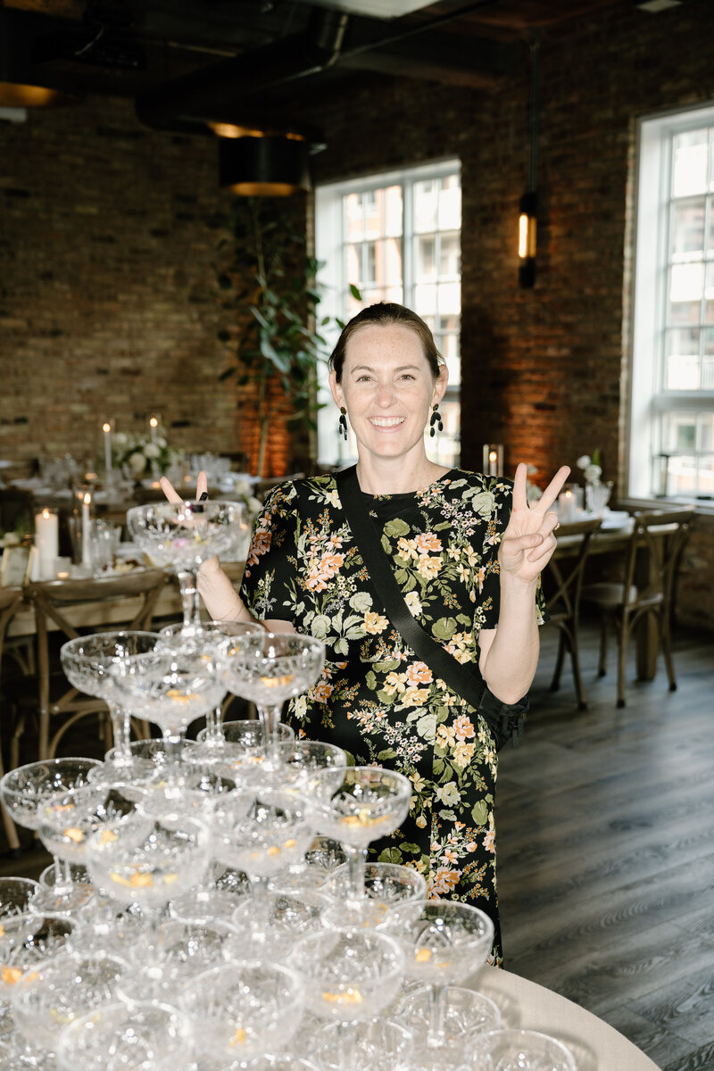 Bridget wears a floral dress with her hair pulled back and holds up two peace signs on a client's wedding day standing behind a table of stacked cocktail glasses.