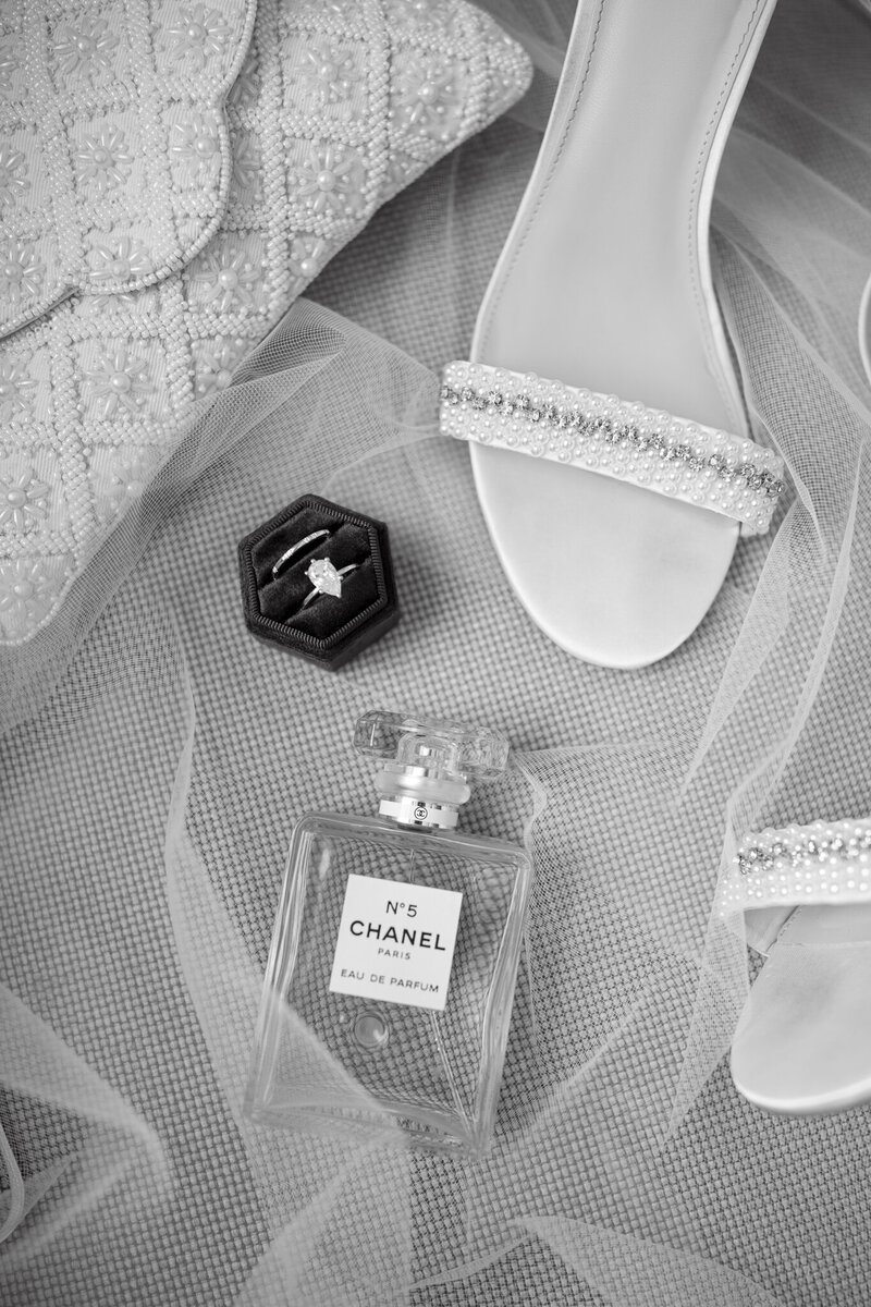 Black and white flat lay of bridal details with Chanel No. 5 perfume, heels, ring, and veil
