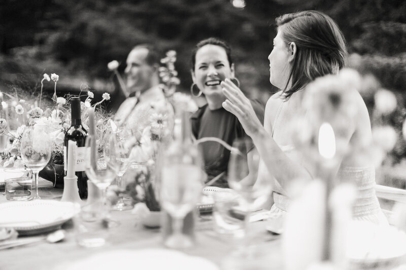 A black and white image of friends laughing at a rehearsal dinner table in the Adirondack Mountains