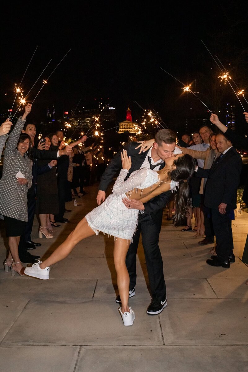 A joyful couple shares a dramatic dip-and-kiss at the center of a sparkler tunnel, surrounded by cheering guests. The bride wears a white beaded mini dress and sneakers, her leg extended in a dancer's pose, while the groom leans in with a wide smile. The warm glow of sparklers lights the night sky, illuminating the celebration in motion and the thrill of a just-married moment.