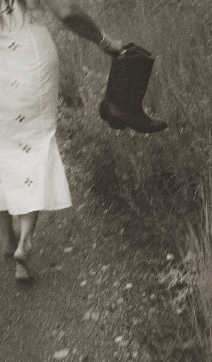 Aesthetic photo of girl in white dress holding cowboy boots walking through field