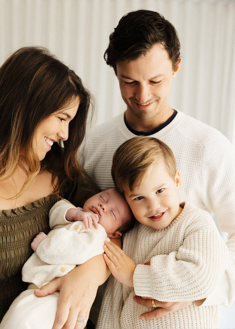 A mother and father hold their newborn baby and a little boy while the boy grins at the camera and the parents look at their children and lucy takes photographs of them in her newborn photography studio
