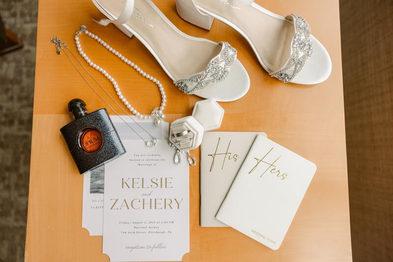 Wedding flat lay details with jewelry, heels, stationary, and vow booklets.