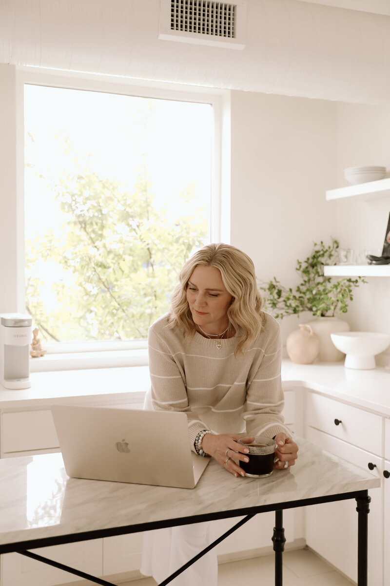 A woman sitting in a kitchen on her computer looking up at the camera