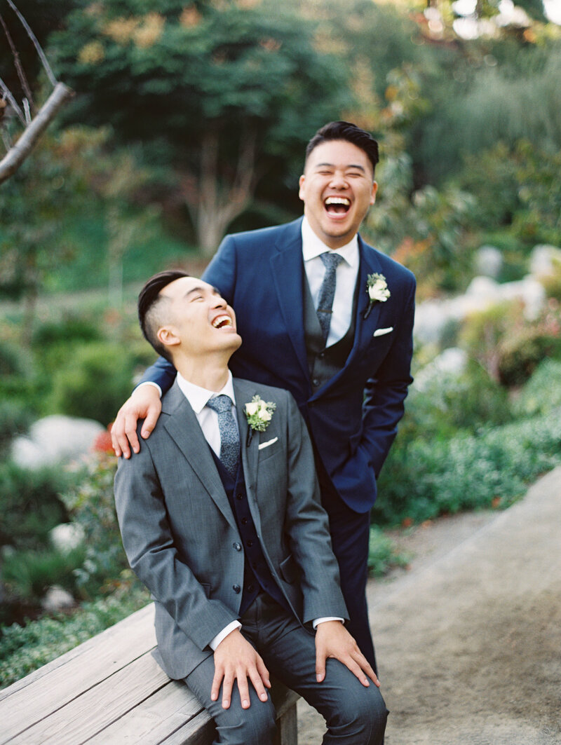 Two grooms, one sitting, and the other standing, laughing out loud at the Japanese Friendship Garden in Balboa Park