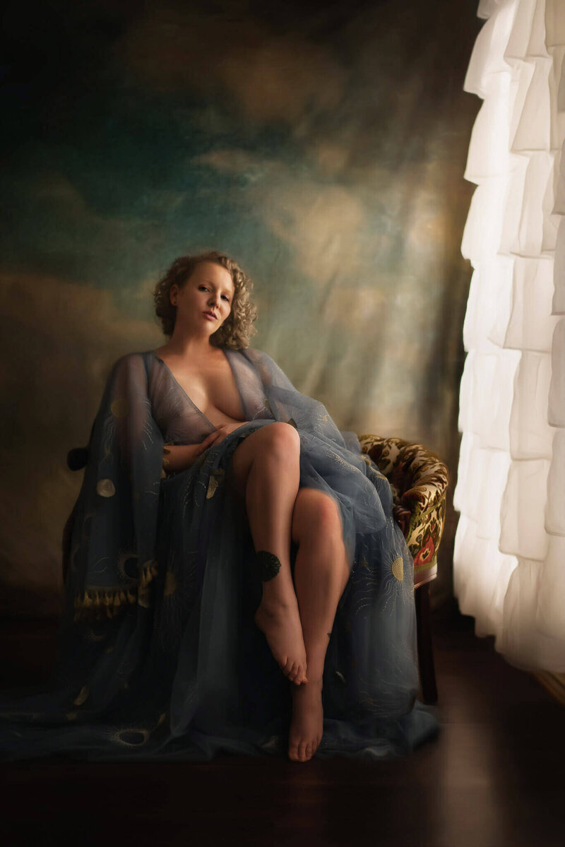 Woman in a sheer blue gown sitting on a bench in front of a cloud backdrop in a DFW boudoir studio