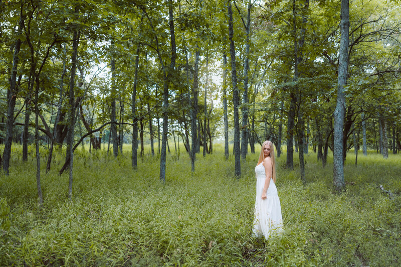 High school senior standing in green grass in a forest wearing a long flowy dress. 