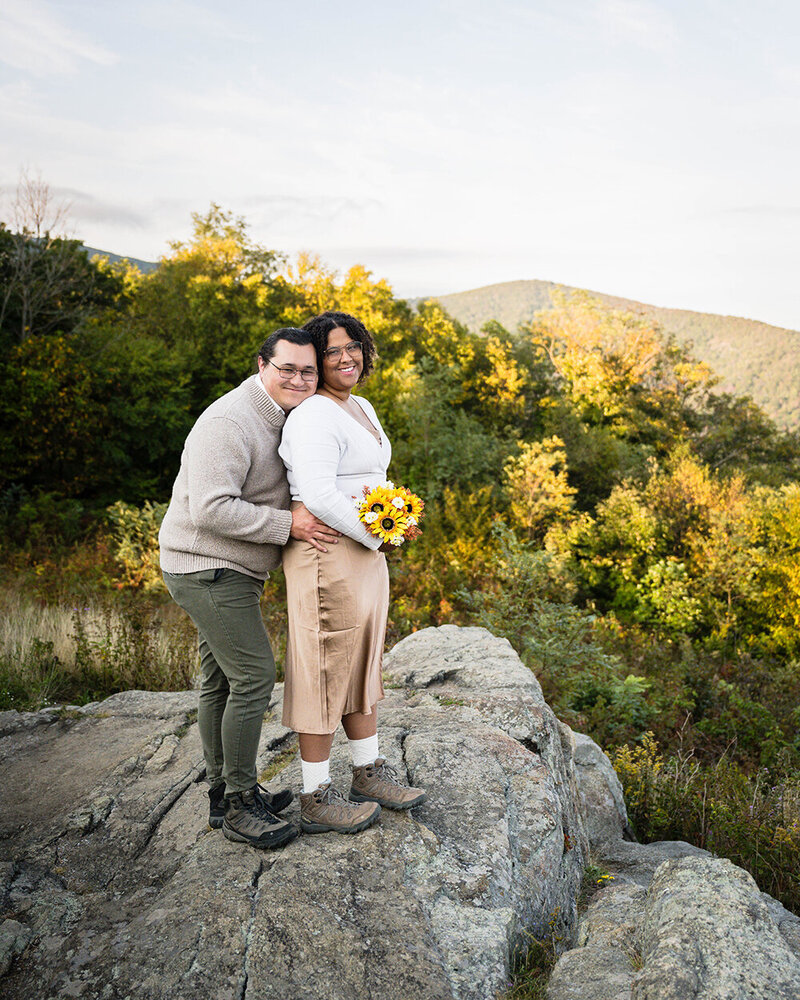 A couple on their elopement day stand on a large rock overlooking the mountains on their elopement wedding day in Shenandoah National Park in Virginia.
