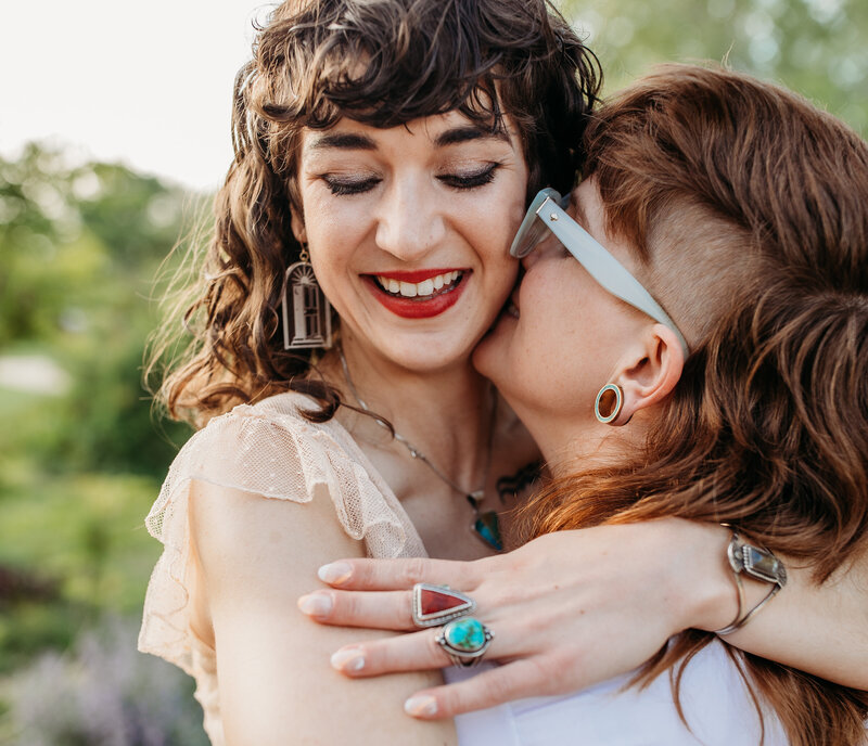 A woman with curly hair and vintage earrings smiles as another person whispers into her ear