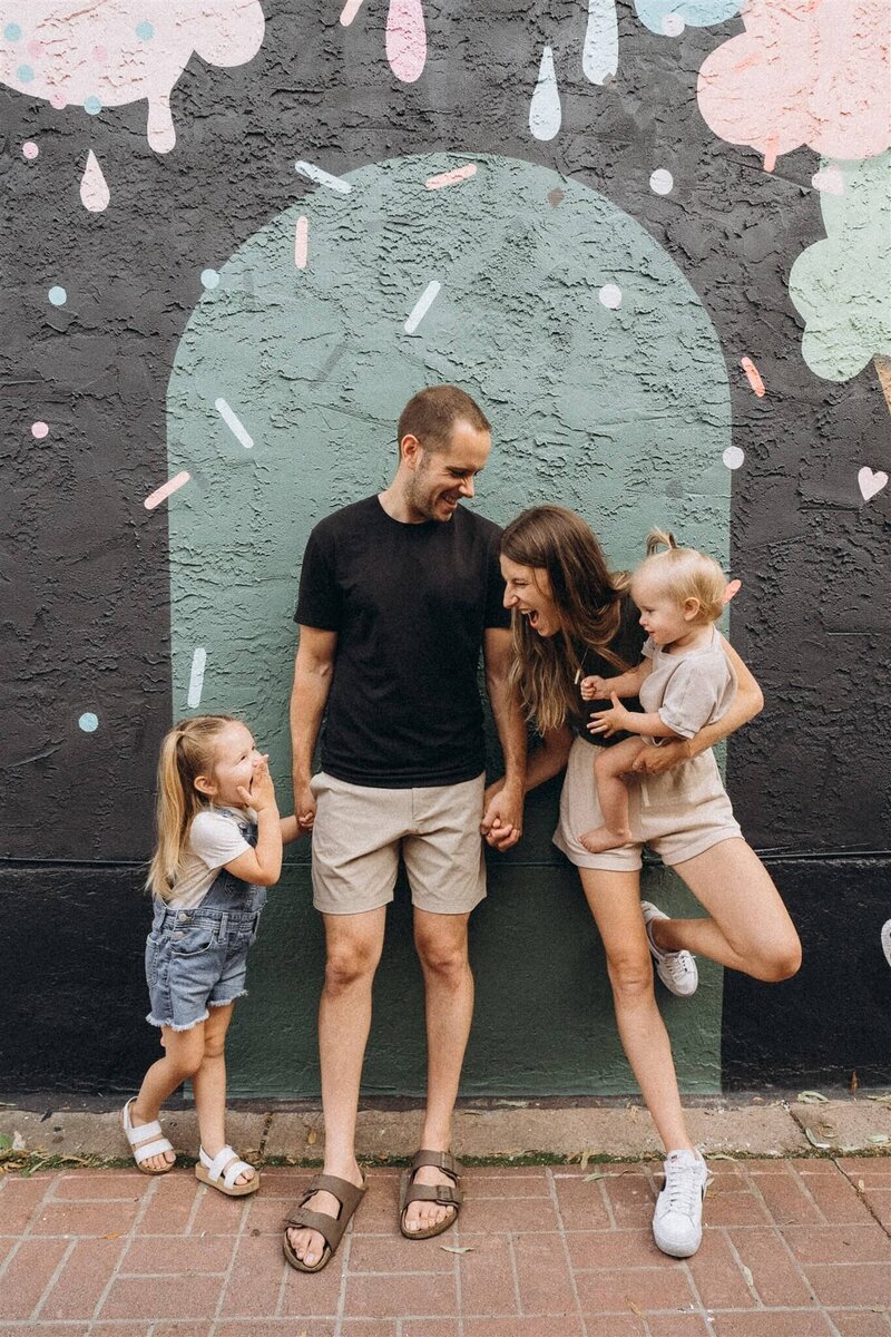 Cinematic wedding photographer based in Minnesota and Florida laughs with her daughters and husband as they pose in front of ice cream mural wall