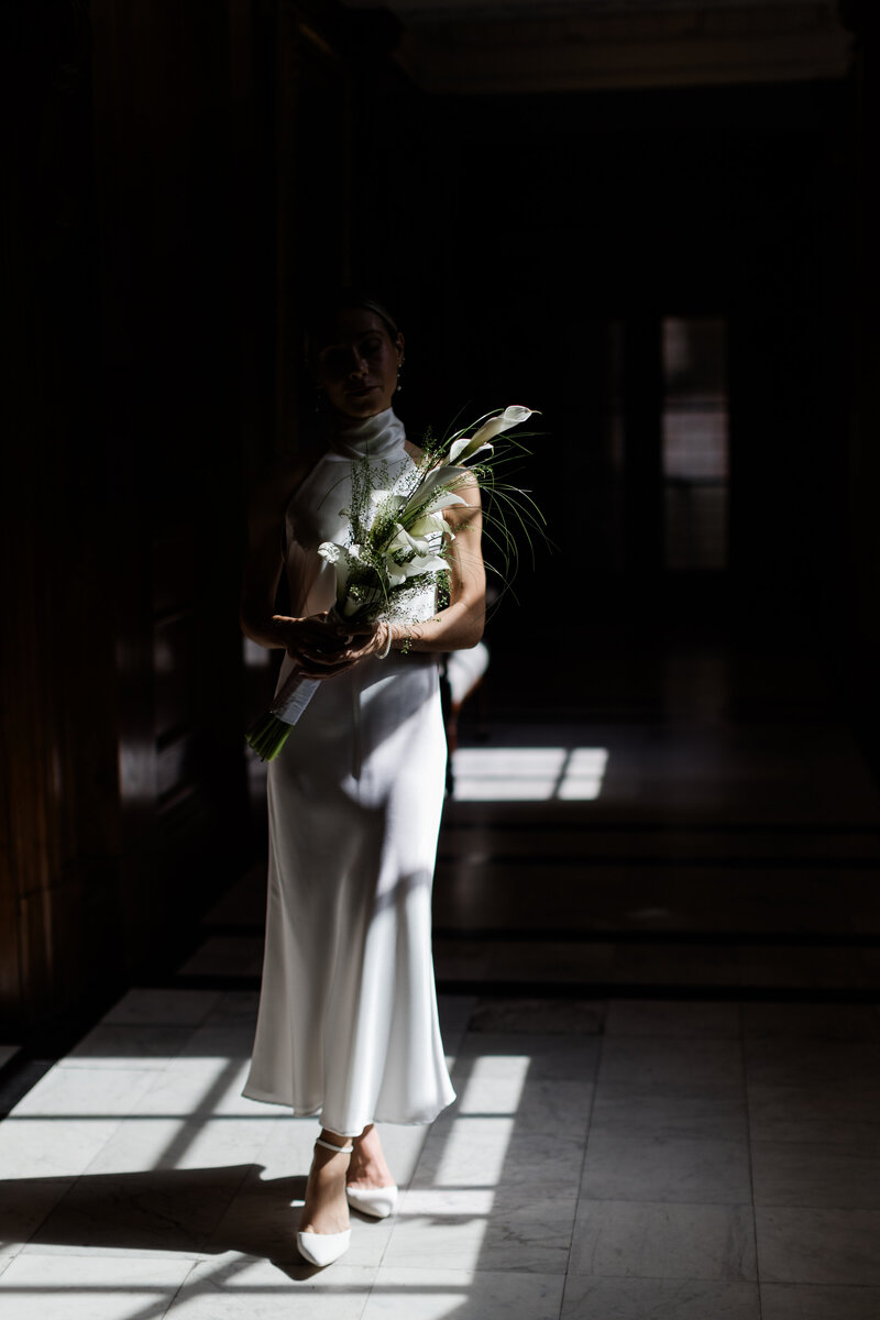 Bride in Emmy bridal shoes standing in pocket of light in the corridor of Old Marylebone Town Hall holding a bouquet of long stem calla lilys