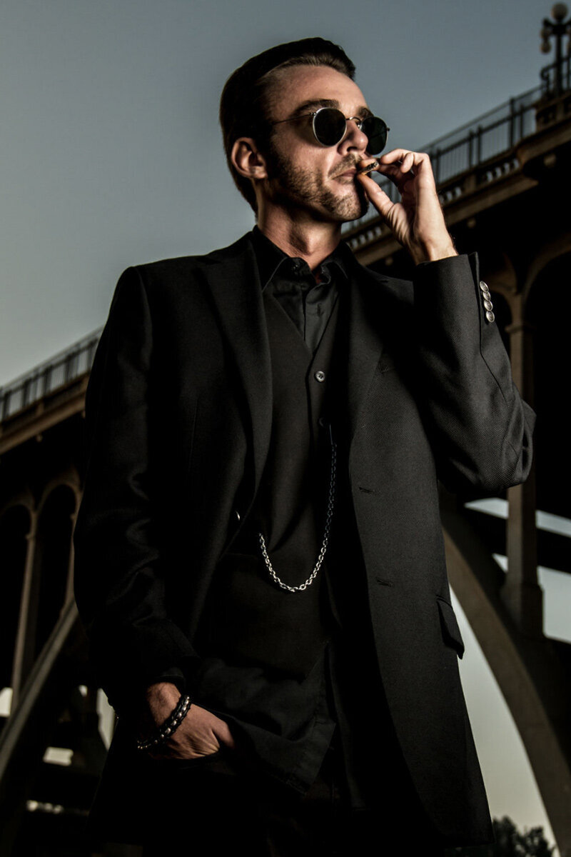 Professional Branding Portrait Author Chandler Morrison standing beneath bridge while smoking wearing black suit with silver pocket watch chain wearing sunglasses
