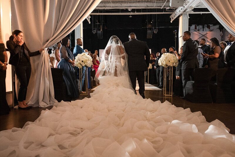 Bride walking down the aisle at The Crane Bay Event Center in Indianapolis, wearing a custom lace wedding dress with a long ruffled train, surrounded by elegant white floral arrangements and modern drapery during a luxury indoor wedding ceremony.

