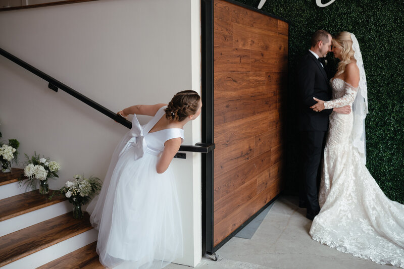 Bride and groom kiss in a dark corner while a young girl in a white dress peeks at them around the corner of a wall