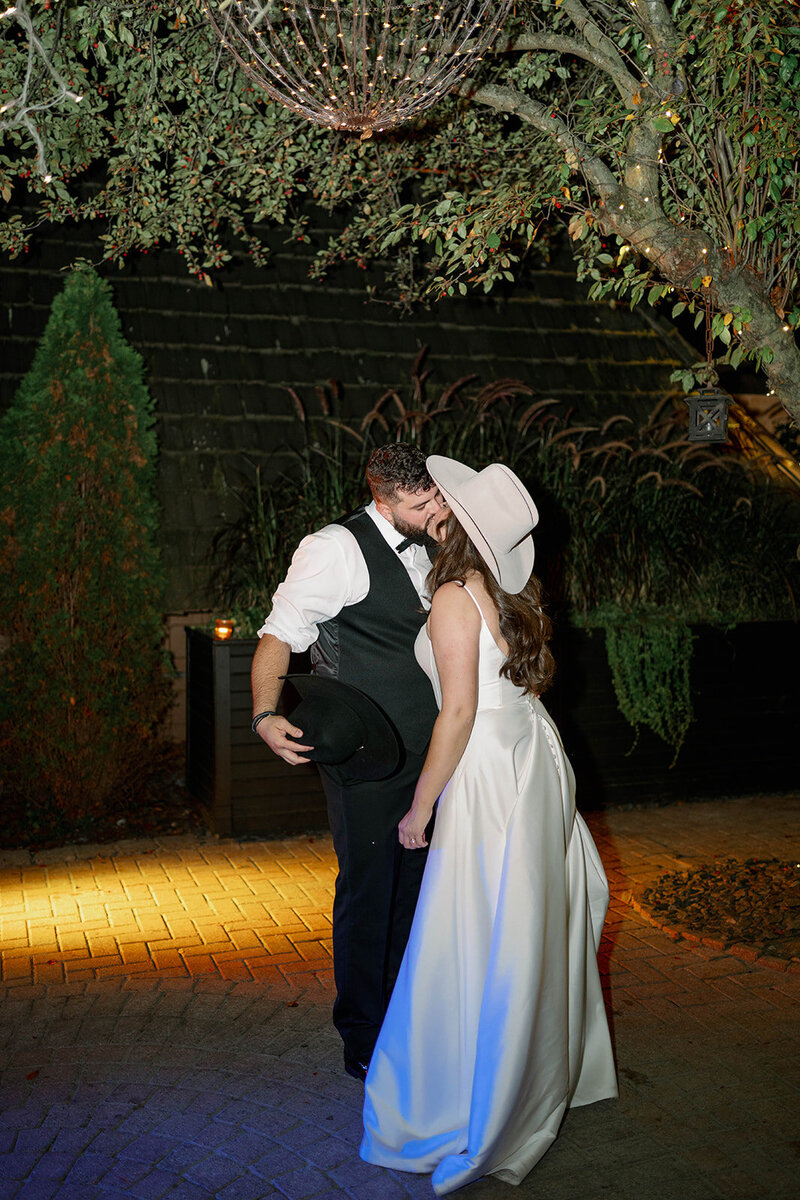 Bride and groom sharing a kiss on the outdoor dance floor at Café Cortina, wearing cowgirl and cowboy hats during their wedding reception.