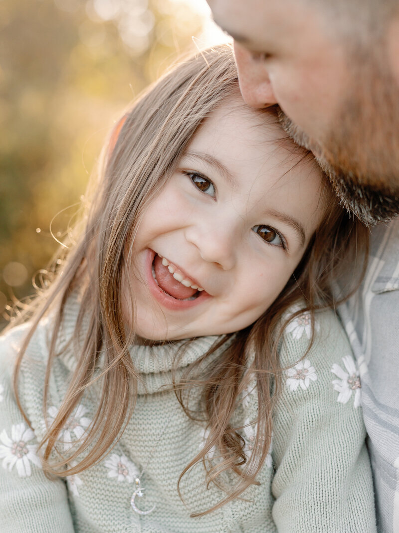 A sweet young girl with bright eyes and a radiant smile tilts her head playfully, bathed in warm natural light. Her carefree expression captures the beauty of childhood in a candid and timeless family portrait. Photographed by KCP Families.