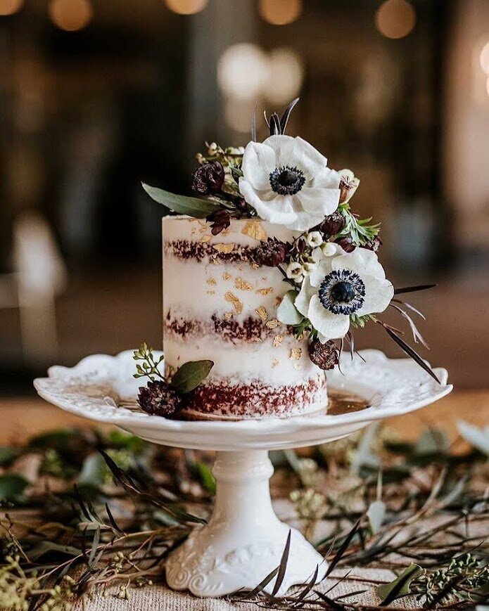 Photo shows a small cake on a white pedestal cake stand, surrounded by greenery on the table. The cake is a naked-style red velvet cake, with white anemone flowers, purple accent flowers, and gold leaf scattering. 
