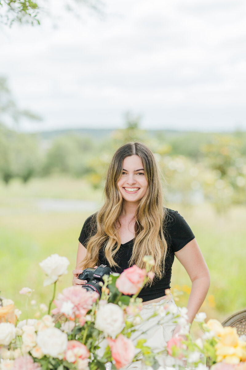 New England Wedding Photographer Liana Huot is captured standing behind a wedding tablescape. She is smiling at the camera with her own camera in hand.
