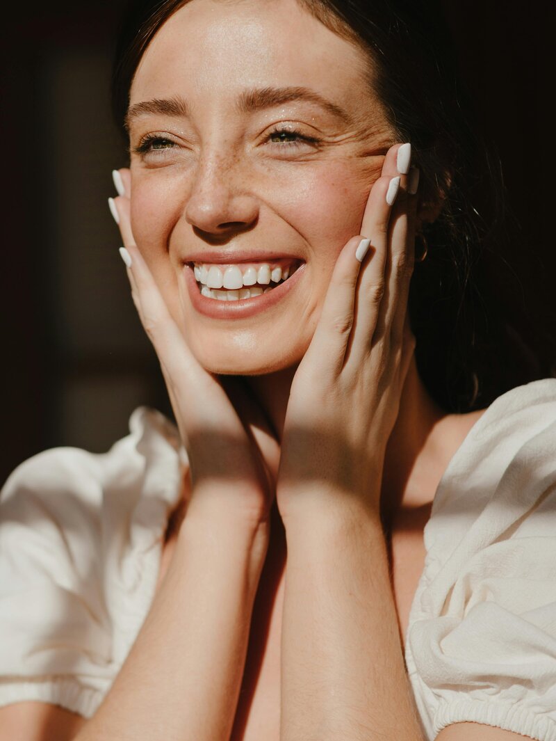 Side-by-side portraits of a man and a woman. The woman, indoors, is smiling broadly with her hands on her cheeks, natural light illuminating her face.