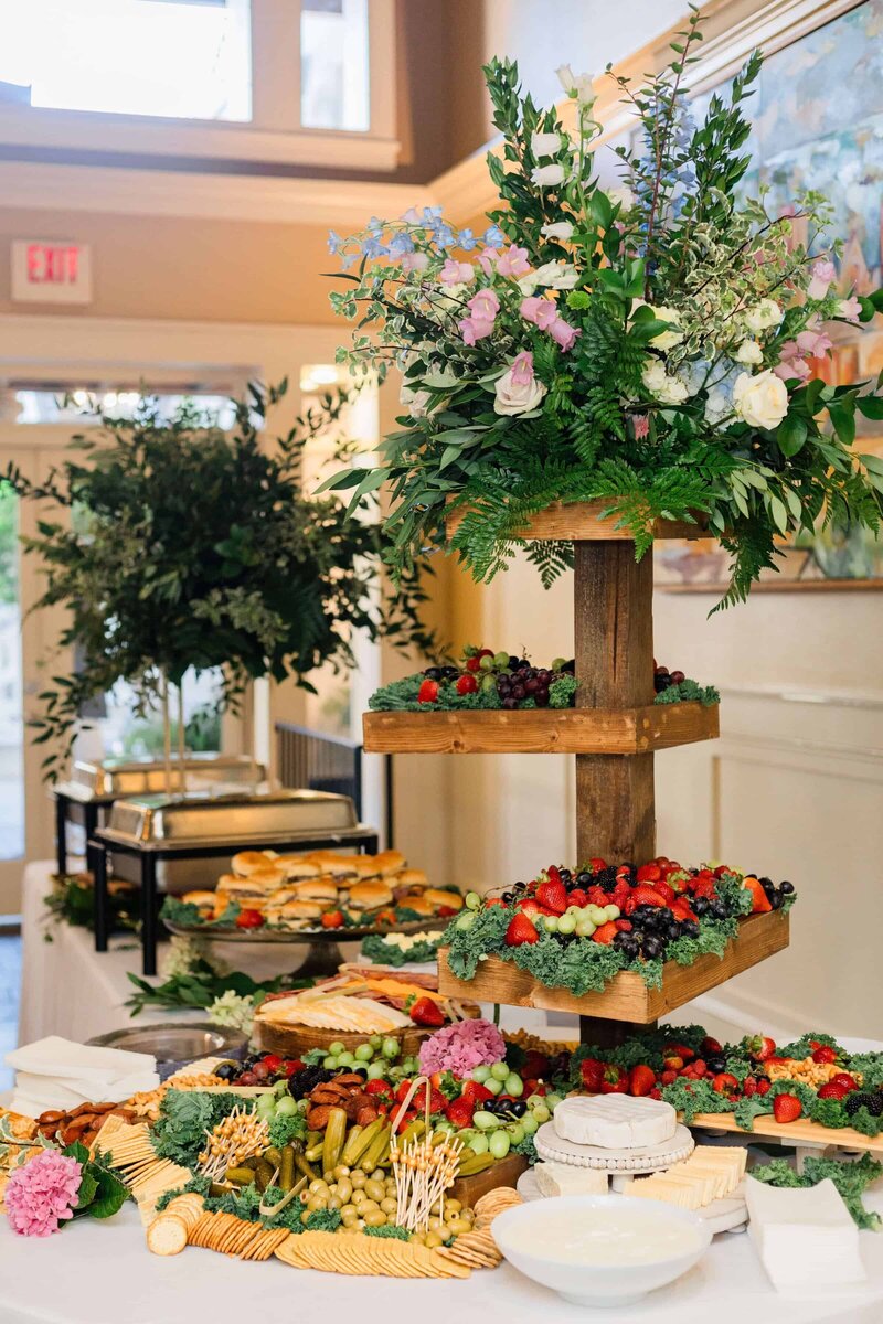 A buffet table with a wooden tiered stand holding assorted fresh fruits and leafy floral arrangements, surrounded by cheese, crackers, vegetables, and a tray of sandwiches in a bright, elegant room.