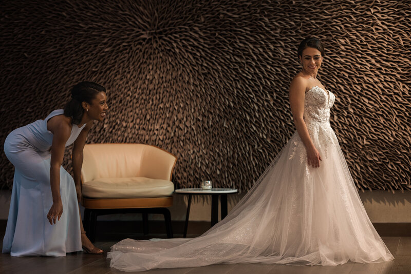 female black woman in a dusty blue chiffon gown smiling and fluffing the train of a white brunette bride with a white strapless full length ballroom gown smiling back at the black female in front of a brown textured backdrop
