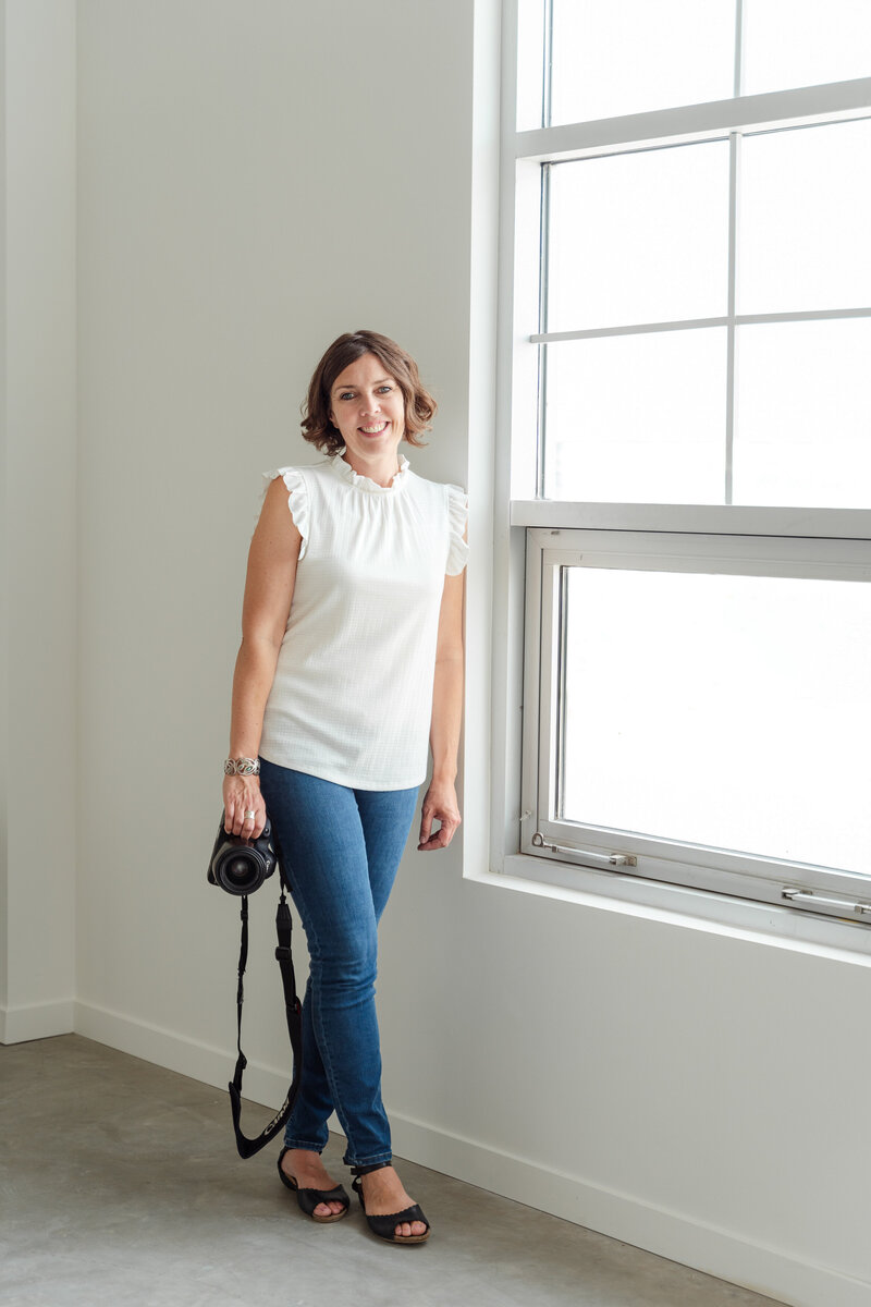Calgary Brand Photographer Kate Daniel Photography, smiling, in a white room, standing beside a window while holding her camera. 