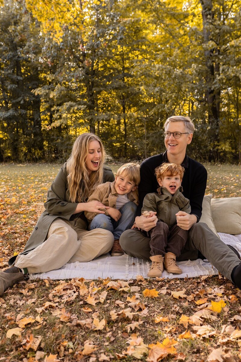 family of four sit on a white blanket surrounded by fall leaves, parents tickling and laughing with their sons