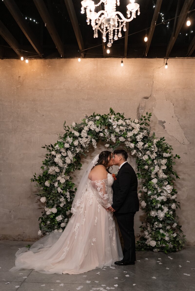 Bride and groom kiss in front of a white floral arch at their Ivory Foundry wedding in Indianapolis