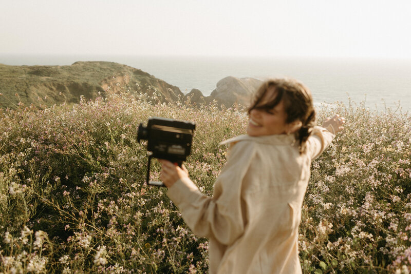 Alexandra Eveland holding her super8 camera, smiling, and standing in front of the san francsisco coast