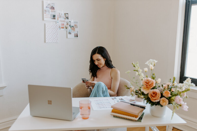 Woman with dark hair and pink tak top and jeans sitting at white desk on her phone and laptop