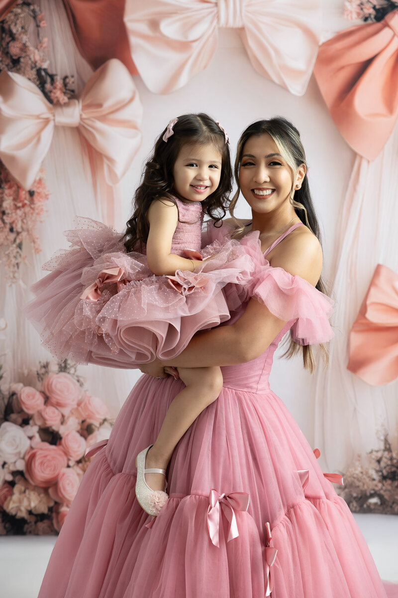 Mother holding her toddler daughter while in matching gowns, smiling during their photography session at LV Photo Haus
