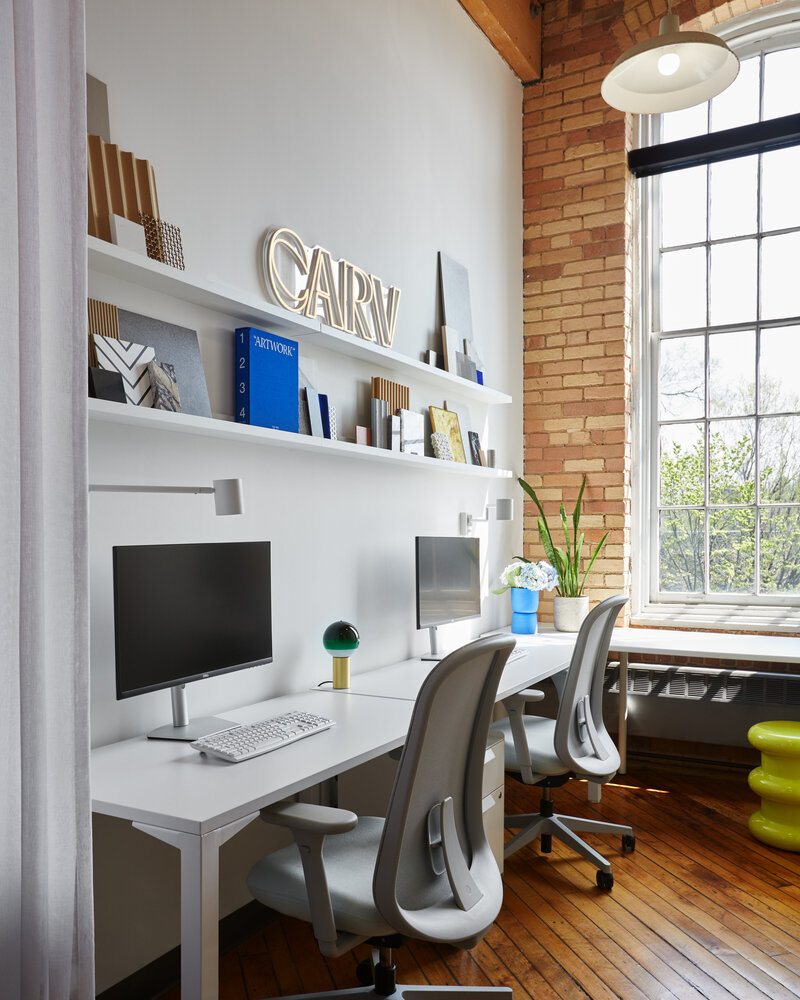 Two white chairs and desks with computer monitors positioned against a wall with floating shelves containing various books, art pieces, and CARV's logo. A tall paned window surrounded by brickwork and two potted plants are positioned adjacent to the desks.