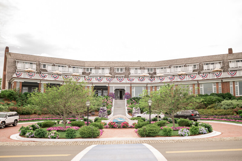 A wide view of the front entrance to Chatham Bars Inn, decorated with patriotic bunting and lush summer landscaping, showcasing the elegant curved architecture of the historic Cape Cod resort.