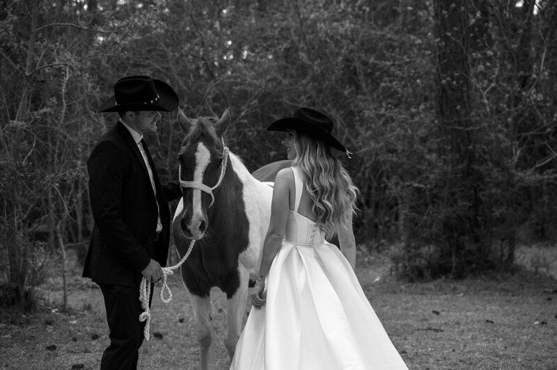 A couple in formal attire and cowboy hats stand with a horse in a forest clearing. The scene is serene, with a rustic, romantic tone.