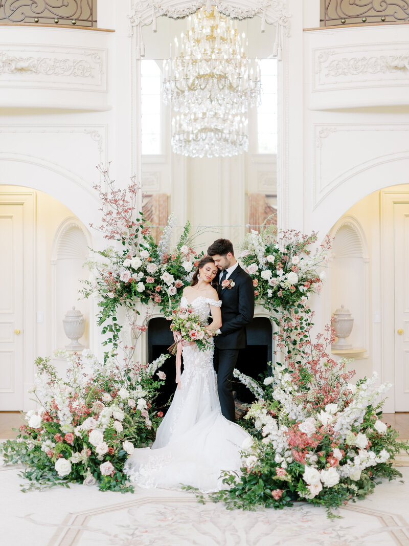 Bride and groom stand against fireplace with overflowing pink, white and green florals at The Olana outside of Dallas, Texas