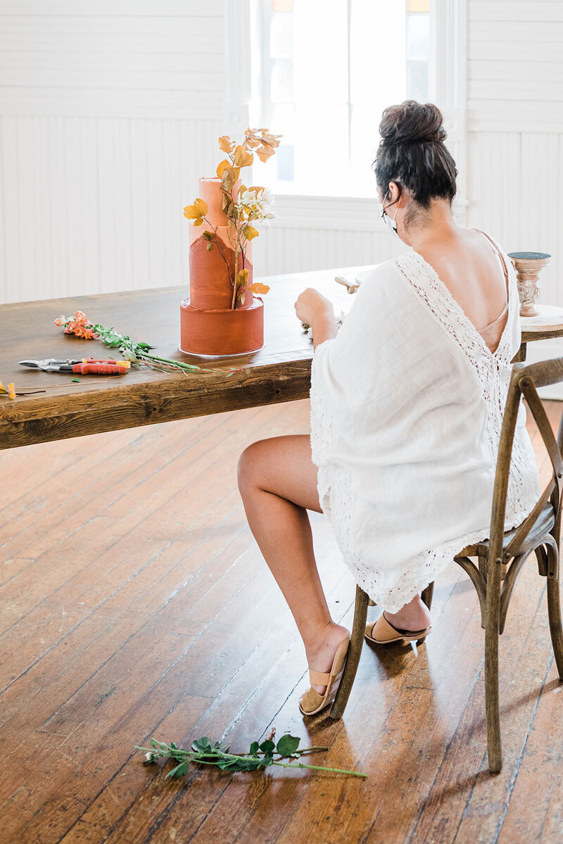 Woman in white dress sits at a wooden table decorating a wedding cake